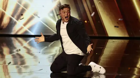 Dymond/TalkbackThames/Shutterstock Harry Moulding on his knees with hands out on the stage as golden paper flies around. He is wearing black jeans and a black jacket with a white T-shirt and white trainers.