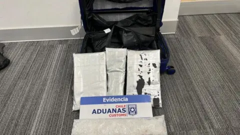 Chile Aduanas Customs A cabin-sized suitcase which has been cut to reveal a secret compartment pictured next to bags of white powder which is methamphetamine. There is a sign by the bags of powder which says 'Evidencia Chile Aduanas Customs'.