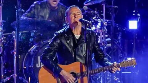 Getty Images Bryan Adams performing onstage in 2022. He is holding an acoustic guitar and standing in front of a microphone. Behind him is a drummer and lots of blue lights. Bryan Adams is wearing a white top and black leather jacket. 