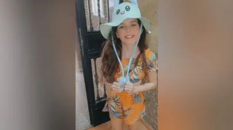 Family handout A young girl with long brown hair wearing matching orange shorts and top with a palm tree design on them and a blue summer hat with a cartoon character face on it. She is smiling up at the camera. She is standing in front of a door.