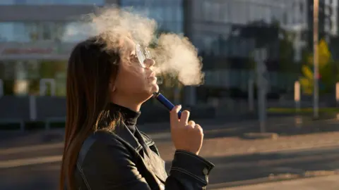 Getty Images A woman with long brown hair and glasses is smoking a disposable vape. There is a puff of white smoke in the air. She is wearing a black jacket and turtle neck as she stands on the street.