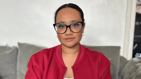 BBC Shanice Aird, who is wearing black-rimmed glasses, a red jacket and has her dark hair tied back, she is sitting on a grey sofa