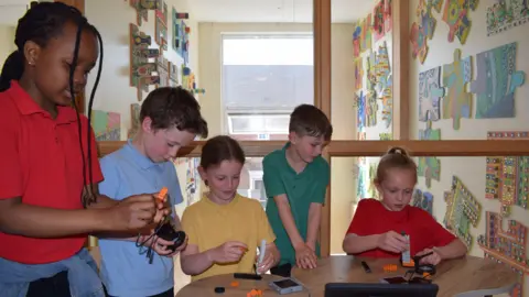 BBC Swindon school children building with Lego style blocks, all wearing bright air tech style tops. Two children are wearing red, one is wearing yellow, one is in blue and one in green. They are gathered around a central table all focussed on what they are building. In the background, there is a wall with big puzzle piece as art on it.