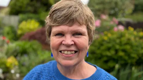 BBC Gwen, who has short blonde hair, is looking at the camera and smiling. She's wearing a blue crew-neck jumper and she has blue earrings. She's standing in a garden which is out of focus behind her. There are many green plants and bushes.