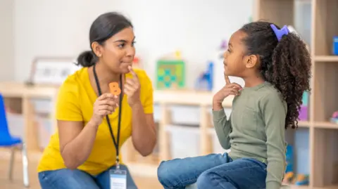 Getty Images A female therapist with dark hair sits with a little girl during a speech therapy session. They are using colourful letters as the therapist holds them up one by one and works through the sounds with the little girl.