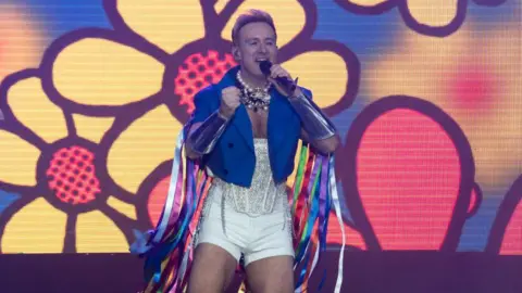 Getty Images Ian Watkins on stage with yellow flowers projected on screen behind him wearing white shorts and strapless sequinned top and cropped blue jacket with coloured ribbons flowing down from the arms