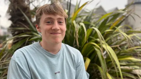 BBC Jacob Mallon is wearing a blue t-shirt while sitting on a wall. He is sat in front of a large green palm.