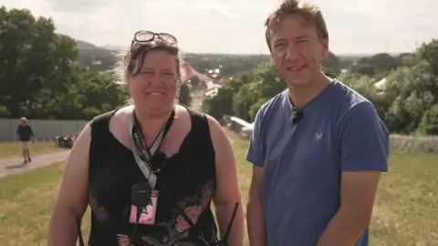 BBC Gareth Churcher and Jade Dunbar standing outdoors with a scenic background of trees and a distant view of a town or festival. Jade Dunbar wears sunglasses on their head, a black sleeveless top, and a lanyard. Gareth Churcher wears a blue t-shirt.