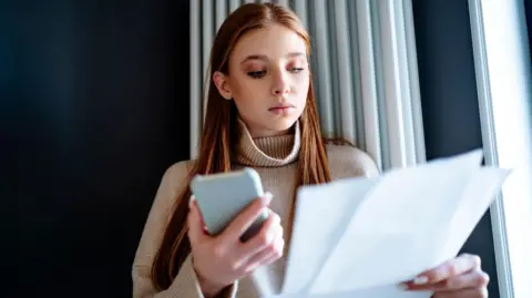 Getty Images File photo of a woman with long brown hair looking at some papers (probably bills) which she is holding with her left hand. She holds a mobile phone in her right hand. She is wearing a light-coloured top with a high neck and standing in front of some light-coloured curtains.