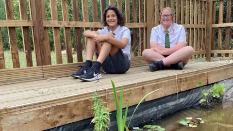 BBC Siena and Alazar sit on some wooden decking with a pond in front of them. They are both smiling wearing white shirts, ties and grey shorts.