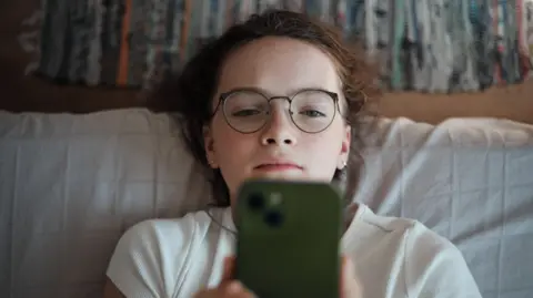 Getty Images A young teenage girl looks at a smartphone. She is wearing glasses and a white T-shirt and leaning against white pillows or cushions with coloured curtains in the background.