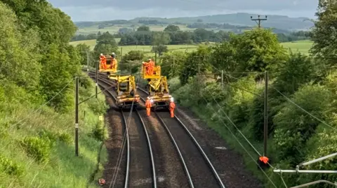 Network Rail Scotland Network Rail staff wearing orange suits and hard hats work to repair the overhead train lines at Beattock.