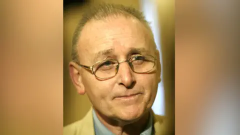 Pacemaker A close up image of Denis Donaldson. He has thinning, short grey hair and has a neutral expression on his face as he takes a question at a press conference. He has a pair of silver glasses on. He is wearing a tan suit jacket and a blue collared shirt.