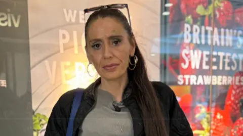BBC Keeley is stood outside some shop windows. She has her hair tied back and sunglasses on her head and is wearing a black jacket and grey top.