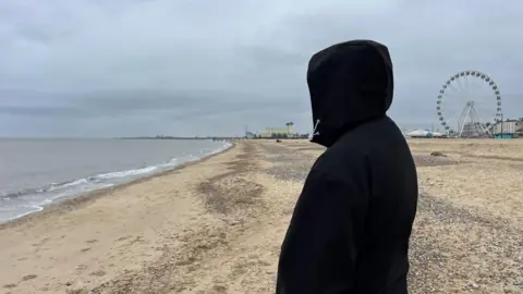 Andrew Turner/BBC A picture of Leon in a black hoodie, with the hood covering his face to protect his identity. He is pictured on Great Yarmouth's central beach, with the Wellington Pier and big wheel visible in the distance.