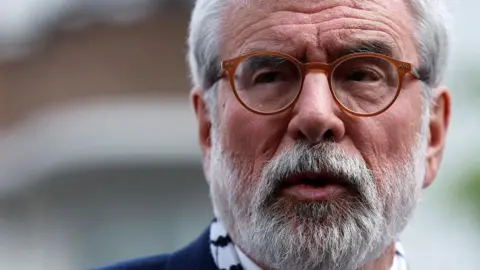 Reuters Gerry Adams has short white/grey hair and beard. He is wearing brown/orangey framed glasses and is speaking outside court in Dublin.