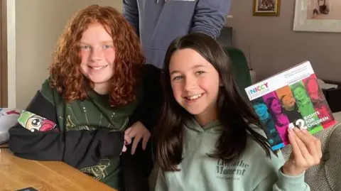 NICCY Astrid Knox has red curly hair and is wearing a a black and green hoody. Rhea Flood has long dark hair and is wearing a sage green hoody. They are sitting at a kitchen table holding a children's commissioner report entitled 12 rights for children.