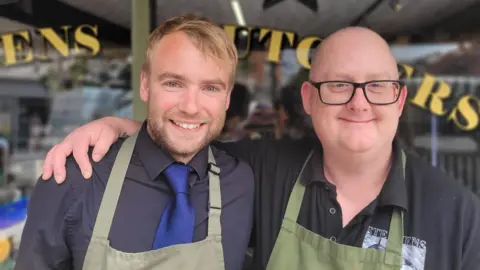 BBC Two men are standing in front of a window to a butchers with their arms around each other. Both are wearing black shirts and green aprons.