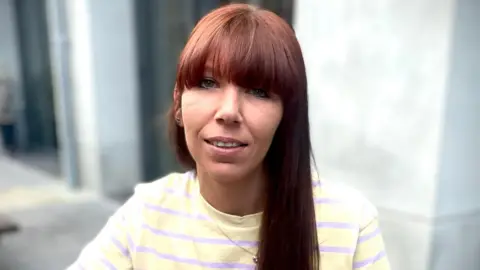 BBC A woman with long reddish-brown hair looks into the camera. She is sitting outside. We can see her from the chest up. She is wearing a yellow t-shirt with light purple stripes and has a chain necklace. The background is blurred but we can see the wall of a building.