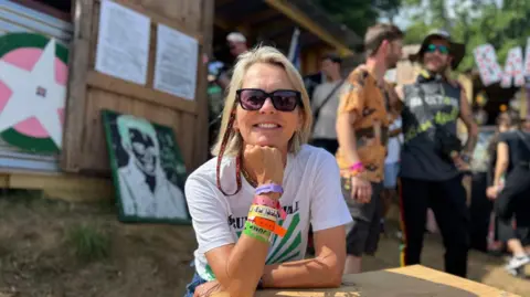 BBC Lucinda Tait sat at a table in Strummerville, wearing sunglasses, a white T-shirt with green stripes and colourful festival wristbands.