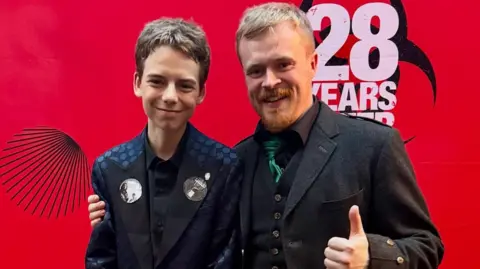 RuMac Alfie, wearing a midnight blue jacket, and Ruairidh, who is wearing a Highland dress jacket, are smiling as they stand in front of a red background with the words "28 Years Later" on it.