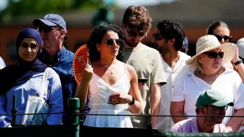 PA Several people standing in a crowd at Wimbledon in the heat. They are dressed in summer clothes, often in caps and sunhats. The woman in the centre of the picture is using a fan and wearing sunglasses.