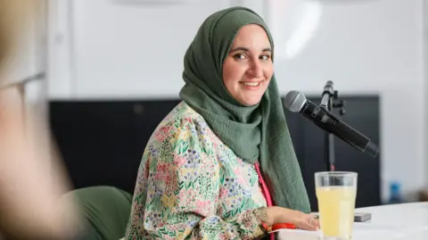Sam Hardwick/Hay Festival Hanan sat at a table with a microphone in a stand. She is turning to smile at the camera and is wearing a top with flowers on it and a sage green headscarf.