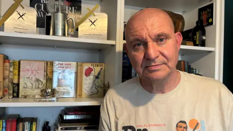 Supplied Mike Craven is standing in front of his bookcase and looking at the camera. He is bald and is wearing a 'Ian Fleming's Dr. No' t-shirt. Two gold dagger awards are displayed, along with some of Fleming's books.