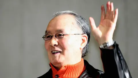 Getty Images A photo of Shigeo Nagashima wearing a orange turtle neck jumper and a black leather jacket. He is waving. 