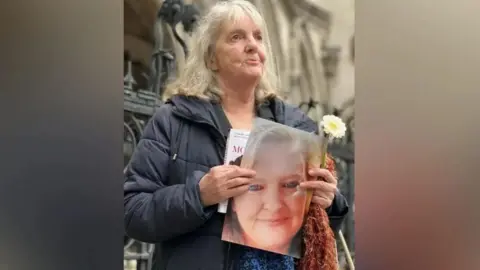 BBC Joy Dove outside a court holding a picture of her smiling daughter. In her left hand she is holding a white flower. 