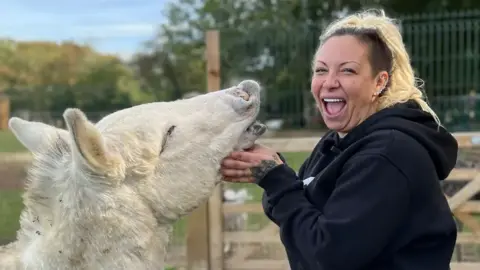 Lauren Carter/BBC Jodie Marsh is standing on the right looking towards the camera smiling. She has blonde hair in a ponytail, and is wearing a black hoodie. She is standing next to a white donkey holding its head.