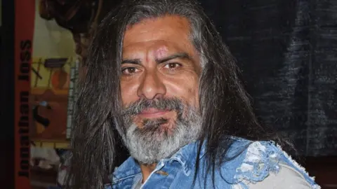 Alamy Jonathan Joss smiling for the camera with long black hair and a frayed denim shirt, pictured in 2021