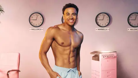 Shutterstock Kyle Ashman poses in pale blue swimming trunks for a Love Island promotional shot. He smiles at the camera, his hands in his pockets, and his image is super imposed on an office-style background complete with pink desk chair and photo copier with four clocks on the wall showing the time in Spanish destinations as just after 9.