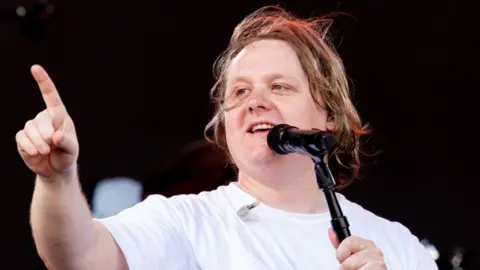 Getty Images Lewis Capaldi on stage at Glastonbury 2023 in a white T-shirt singing into a microphone with one finger pointing in the air