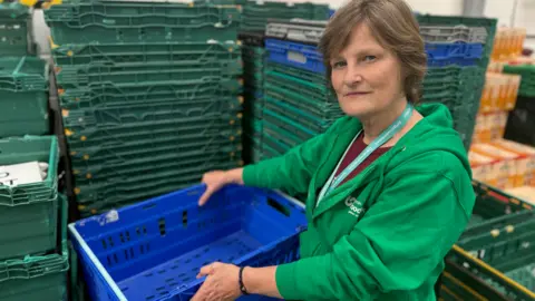 BBC The photo shows a lady with a neutral expression looking at the camera. She is wearing a green hoodie, and is holding an empty blue crate. Behind her are stacks of crates about 5ft (1.5m) high which are also all empty.