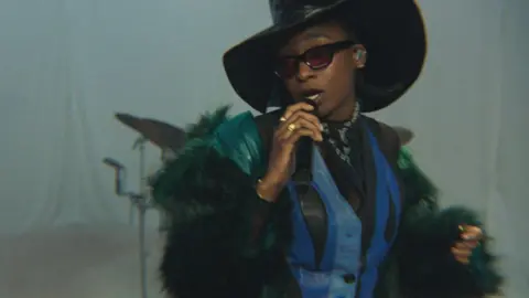 BBC Little Simz performing at the BBC's Maida Vale studios. She wears an oversized leather top hat, blue leather waist coat, green furry coat and dark sunglasses. She raps into a microphone while looking over her right shoulder against a white backdrop.