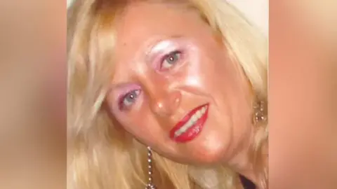 Gardai Tina has blond long wavy hair and blue eyes. She is wearing pink eye shadow and dark eyeliner. She has red lipstick on and long silver dangly earrings. 