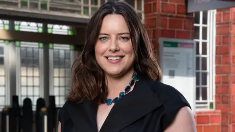 BBC/Jack Barnes Michelle Ryan is smiling on the set of EastEnders, she is stood in front of Walford tube station