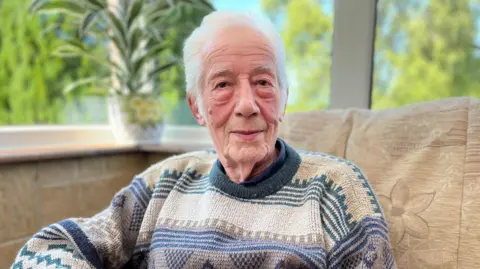 BBC 90-year-old Ernest Williams sat in a conservatory. There are large windows behind him showing blue sky and trees outside. 