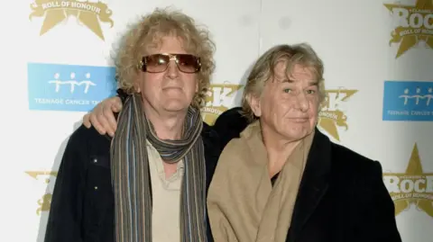 PA Media Ian Hunter (left) and Mick Ralphs (right) arrive for the Classic Rock Roll of Honour Awards in 2006. Hunter has curly blond hair and large sunglasses. He is wearing a striped scarf with a beige t-shirt and a black coat. Ralphs has long, wavy blond hair with a brown scarf and a black coat.