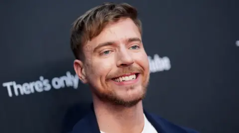 Getty Images MrBeast pictured in New York in May 2025. The creator has blue eyes, short brown hair and a short beard. He smiles, wearing a dark blue blazer over a white T-shirt while posing for photographers in front of black hoarding.