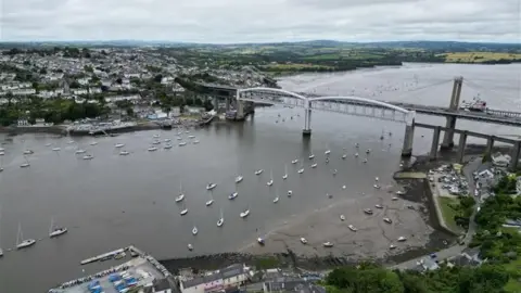 BBC A drone shot of Tamar Bridge. The train bridge and car bridge are visible. There are sailing boats on the river Tamar. Saltash is on the left and Plymouth is on the right. There are pastel houses on both sides. The water is a dark grey. The sky is full of clouds. 