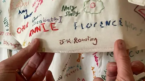 Aleks Kovac A pair of hands are holding a piece of fabric covered in embroided names. The one being held prominently to the camera is "JK Rowling"
