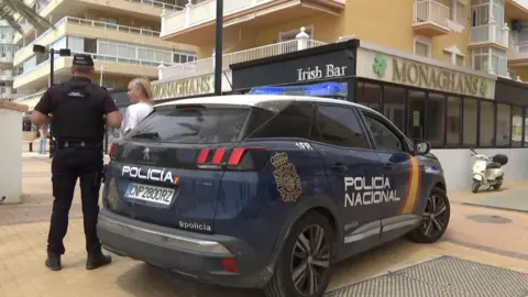 BBC A police car in front of Monaghan's bar. It is blue and there is an officer nearby. He is speaking to a woman.