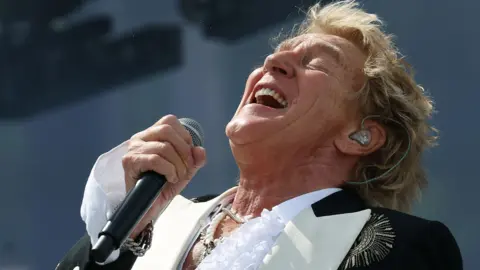 EPA Rod Stewart with a mic in his hand, throwing his head back on the Pyramid stage - he wears a white ruffled shirt and black jacket with gold embellishments 