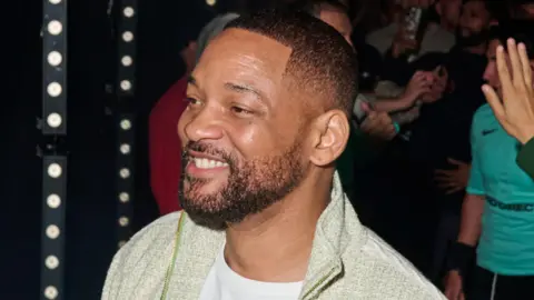 Getty Images Will Smith at Baller League in London. He has a short beard and cropped hair and wears a pale green jumper over a white T-shirt. He smiles, looking away from the camera, with crowds seen behind him in the background. 
