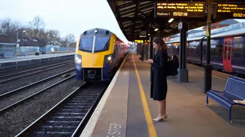 Getty Images A woman is on her phone while she waits on a train platform. There is a yellow and blue train on the tracks in front of her