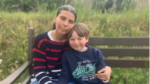 BBC Kim and Henry sit hugging on a wooden bench with long grass seen in the background.