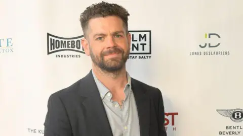 Getty Images Jack Osbourne, who has dark hair cropped short and a beard, standing in front of a hoarding bearing various company logos. He is wearing a grey suit jacket and pale grey shirt and smiling.