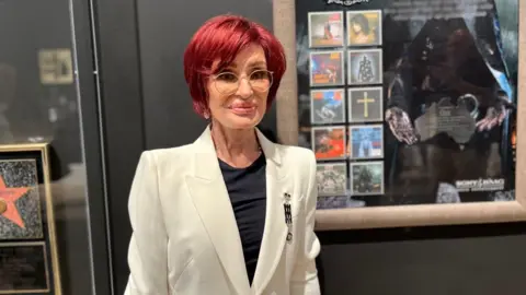 BBC A woman stands in front of framed pictures on a black wall. She has short dyed red hair and glasses. She is wearing a white suit jacket and a black top.